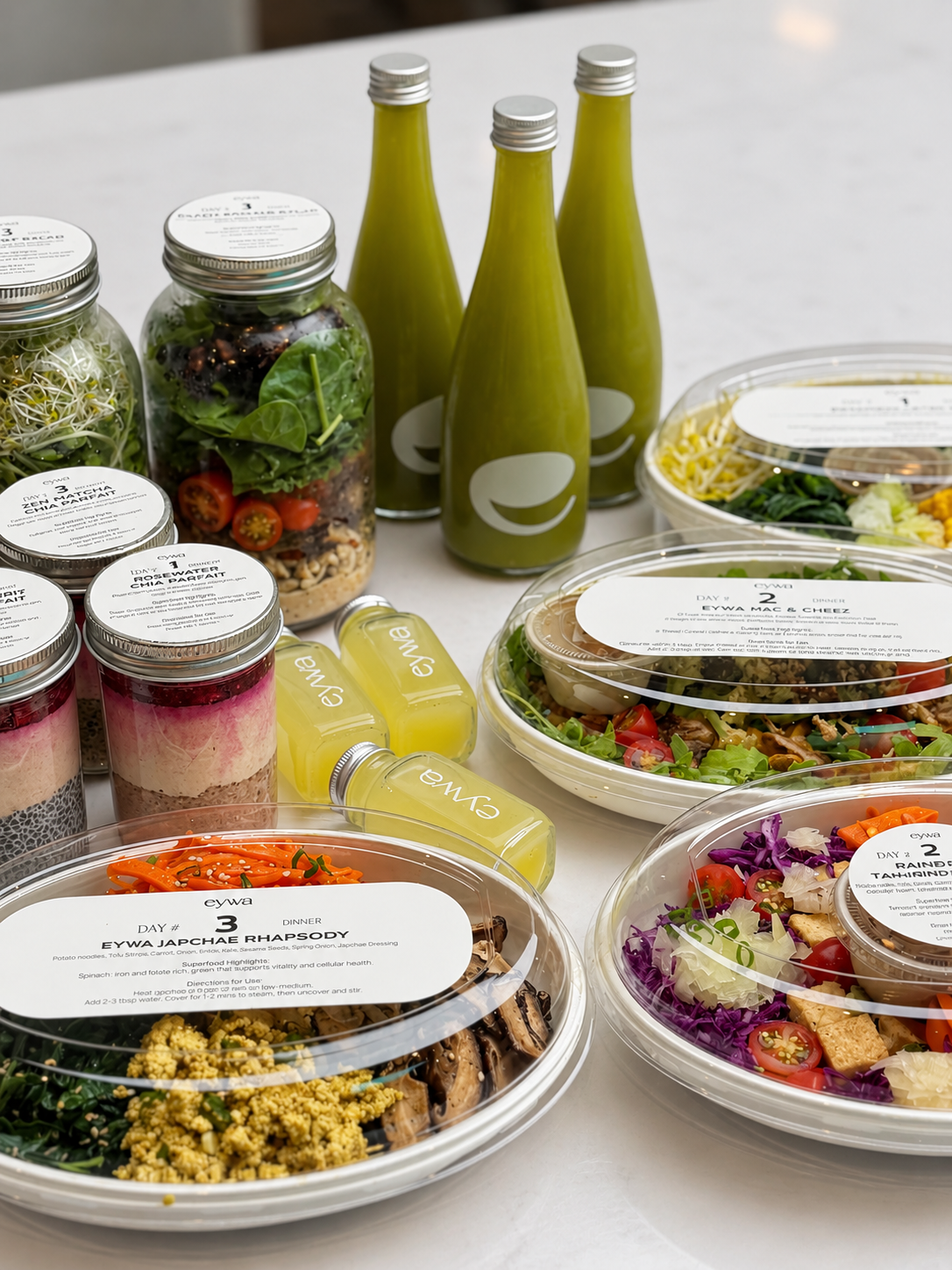 A styled flat-lay of a wellness food and beverage assortment on a white surface, featuring three green drink bottles, several clear mason jars with layered salads and smoothies, and three round plastic bowls of prepared salads.